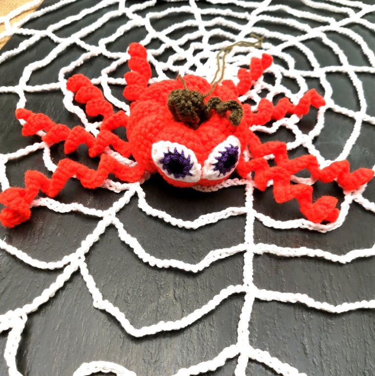 Pumpkin shaped crochet spider with spiral legs & comical eyes sat on a crochet spider web